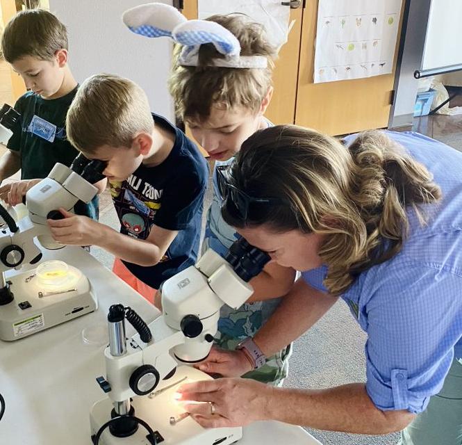 An educator uses a microscope as a child looks on, and two other children use microscopes in the background